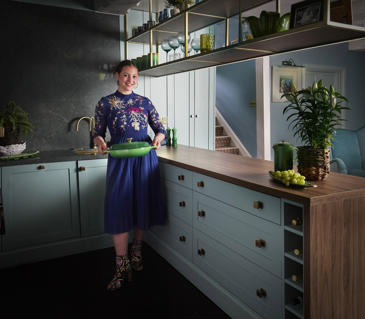 Woman in a blue dress holding a green pan in a teal kitchen.