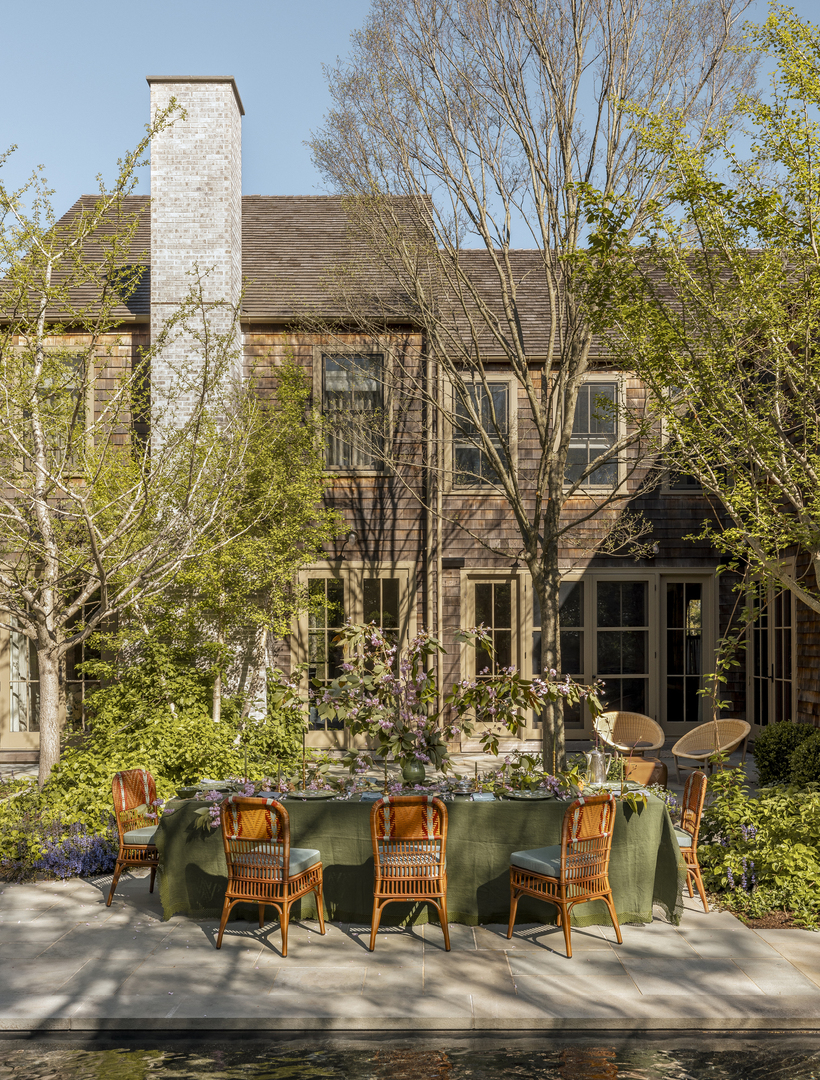 Outdoor dining area with wicker chairs and a green tablecloth, situated in a garden next to a house.